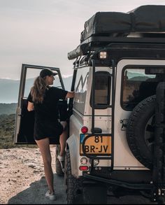 a woman standing next to a white jeep on top of a hill with mountains in the background