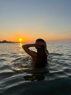 a woman is sitting in the water with her hands behind her head as the sun sets