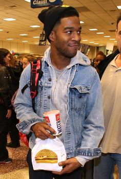 a man in denim jacket and hat holding a bag with a hamburger on it at an airport