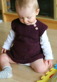 a baby is sitting on the floor playing with a toy train and wearing a knitted dress