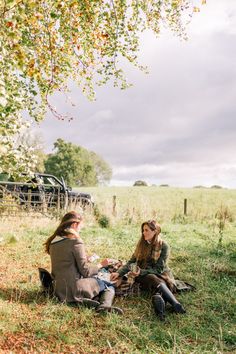 two women sitting on the ground under a tree in an open field, talking and eating