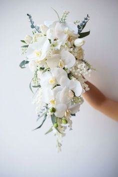 a bridal bouquet with white orchids and greenery is held by a woman's hand