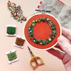 a hand is holding a small embroidery hoop with scissors and spools next to it