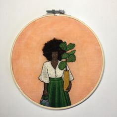 a woman holding a potted plant in front of a cross stitched wall hanging