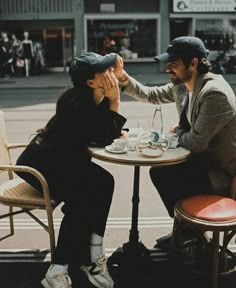 two people sitting at a table with cups and saucers in their hands, one is touching the other's face