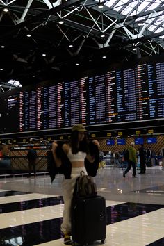 a woman is standing in an airport with her luggage and looking at the departure board