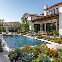 an outdoor swimming pool surrounded by plants and flowers in front of a large white house