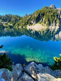 a lake surrounded by trees and rocks with clear water in the foreground, on a sunny day