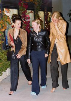 three women are walking down the sidewalk talking on their cell phones while one woman holds her hand up to her ear