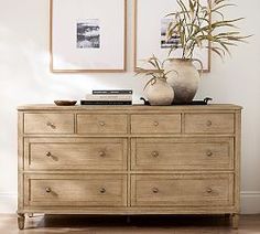 a wooden dresser with two vases on top of it next to a plant and pictures