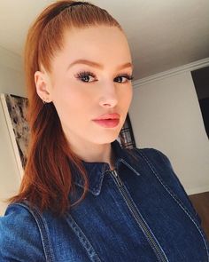 a woman with red hair wearing a denim jacket and looking at the camera while standing in front of a mirror