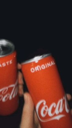 two coca - cola cans are being held up by someone's hands in the dark