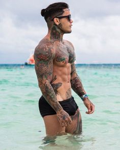 a man with tattoos standing in the ocean looking off into the distance while wearing black swim trunks