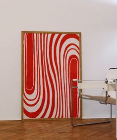 a red and white painting on the wall next to a chair in a room with hard wood flooring