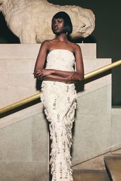 a woman standing in front of a statue wearing a white dress with feathers on it