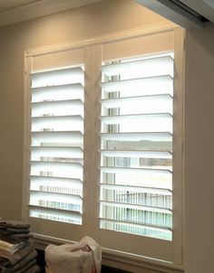 two windows with white blinds in the middle of a room next to a pile of towels