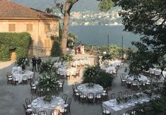 an outdoor dining area with tables and chairs set up for dinner overlooking the water, surrounded by greenery