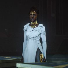 a woman in a white dress with gold accents on her face and shoulders, standing at a stage