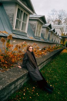 a woman leaning on the edge of a wall in front of a house with autumn leaves