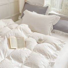 an open book is laying on a bed with white sheets and pillows, along with two gray pillow cases