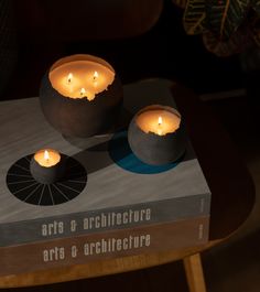 two lit candles sitting on top of a book that reads arts and architecture, in front of a wooden table