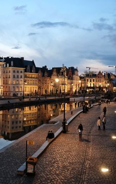 people are walking on the sidewalk near water at dusk in an old european city with cobblestone streets