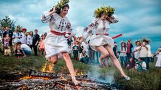 two women in white dresses are jumping over a fire with people watching from the background