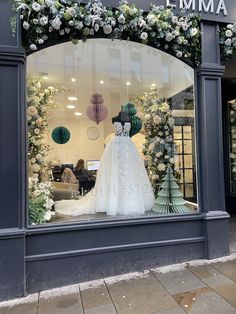 a wedding dress is displayed in the window of a flower shop with white flowers and greenery