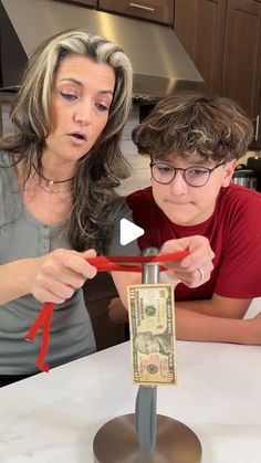 a woman and boy sitting at a table with scissors in front of the dollar bill