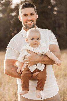 a man holding a baby in his arms and smiling at the camera while standing in a field