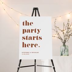 an easel with a sign that says the party starts here on it and string lights in the background