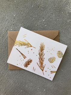 a white card with gold foil on it and some pine cones in the middle, sitting on top of a brown envelope