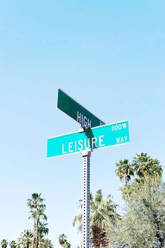 two green street signs sitting on top of a metal pole next to palm tree's