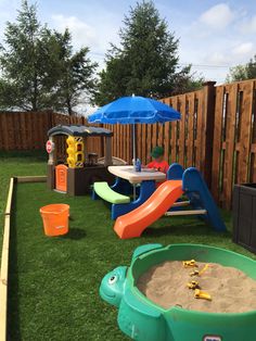 an outdoor play area with sand, toys and umbrellas on the grass in front of a wooden fence