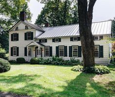 a white house with green shutters and trees in the front yard on a sunny day