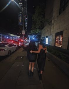 two women walking down the street at night with their backs to each other and cars parked on the road behind them