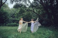 two women in white dresses holding hands and walking through the grass with trees behind them