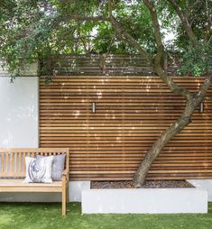 a wooden bench sitting under a tree next to a white wall and green grass covered ground