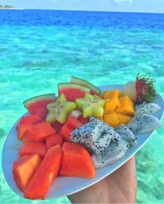 a person holding a plate full of cut up fruit on the beach with blue water in the background