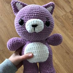 a hand holding a purple crocheted teddy bear with black eyes and white paws