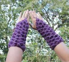 two hands in purple crocheted mitts reaching up into the sky with trees behind them