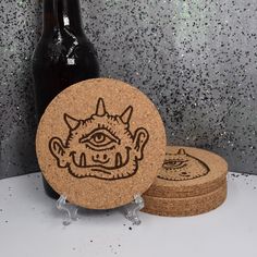 two cork coasters with an image of a demon on them next to a bottle