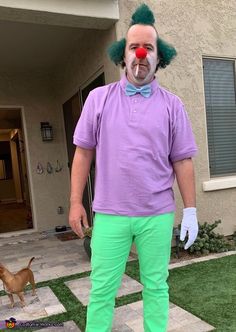 a man dressed as a clown standing in front of a house with a dog on the lawn