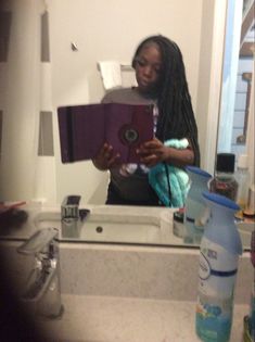 a woman standing in front of a bathroom mirror holding a purple object and looking at her cell phone