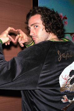 a man with long curly hair wearing a black jacket and holding his hand to his face