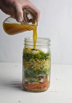 someone pouring dressing into a mason jar filled with vegetables and rice noodles, including broccoli