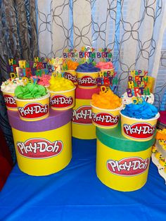 birthday cupcakes in play doh containers are on a blue tablecloth with white curtains behind them