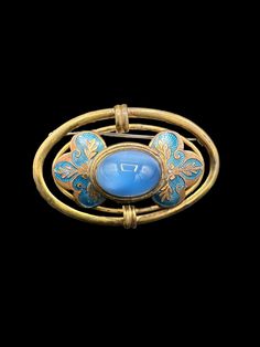 This vintage faux moonstone brooch is stunning. The large oval brass setting, adorned with blue enamel floral decorations, encircles a large sky-blue glass cabochon reminiscent of a moonstone. Its manufacturing date is likely in the 1940s. The glass cabochon could be Czech, but I'm not positive. With its considerable size, the brooch could be a striking scarf pin. While it does show signs of its age with some blemishes to the brass, it still maintains its beauty. To ensure you have a comprehensi Luxury Oval Cabochon Brooch, Blue Oval Brooch Jewelry, Oval Blue Brooch Jewelry, Oval Blue Brooch, Vintage Oval Brooch With Cabochon, Vintage Oval Cabochon Brooch, Blue Oval Brooch, Art Deco Oval Brooch Jewelry, Antique Blue Collectible Brooches