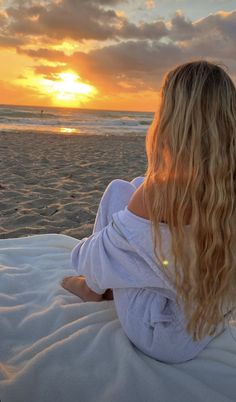 a woman sitting on top of a white blanket next to the ocean at sunset or sunrise