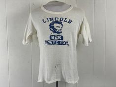 Vintage 1940s school t shirt. Made of white cotton. Has blue Abraham Lincoln graphics. Reads: LINCOLN PHYS. ED. 926. Hand stitched on front label could be removed. Made by WILTON. Size medium. Actual measurements are:   40" around the chest  42" around the waist  25.5" shoulder seam to shoulder seam   24" overall length In good condition with some stains lower front and slightly yellowed towards the top, see pictures. Vintage College T-shirt With Graphic Print, Vintage College T-shirt With Screen Print, Vintage White Top For School, Vintage Fitted Shirt With Screen Print, Classic Fitted School T-shirt, Vintage Screen Print Tops For College, Vintage Cotton T-shirt For College, Retro White T-shirt For School, White Retro T-shirt For School